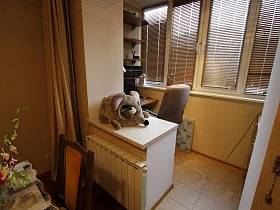 стул у письменного стола с полками на застекленной лоджии с коричневыми жалюзи на окнах ,совмещенной с кухней квартиры государственного служащего с детской комнатой