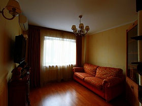 красный цветной мягкий диван с подушками у желтой стены, красные с желтым шторы на окне гостиной трехкомнатной квартиры государственного служащего с детской
