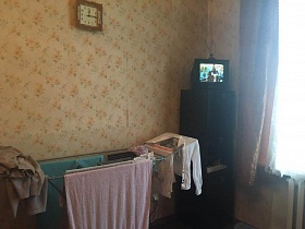маленький телевизор на коричневом узком пенале, белье на сушилке, квадратные часы на стене с цветочными обоями спальной комнаты двухкомнатной квартиры