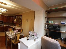 стул у письменного стола с монитором, деревянные открытые полки на светлой стене лоджии, совмещенной с кухней,мягкий домик в виде слоника на столешнице окна трехкомнатной квартиры государственного служащего